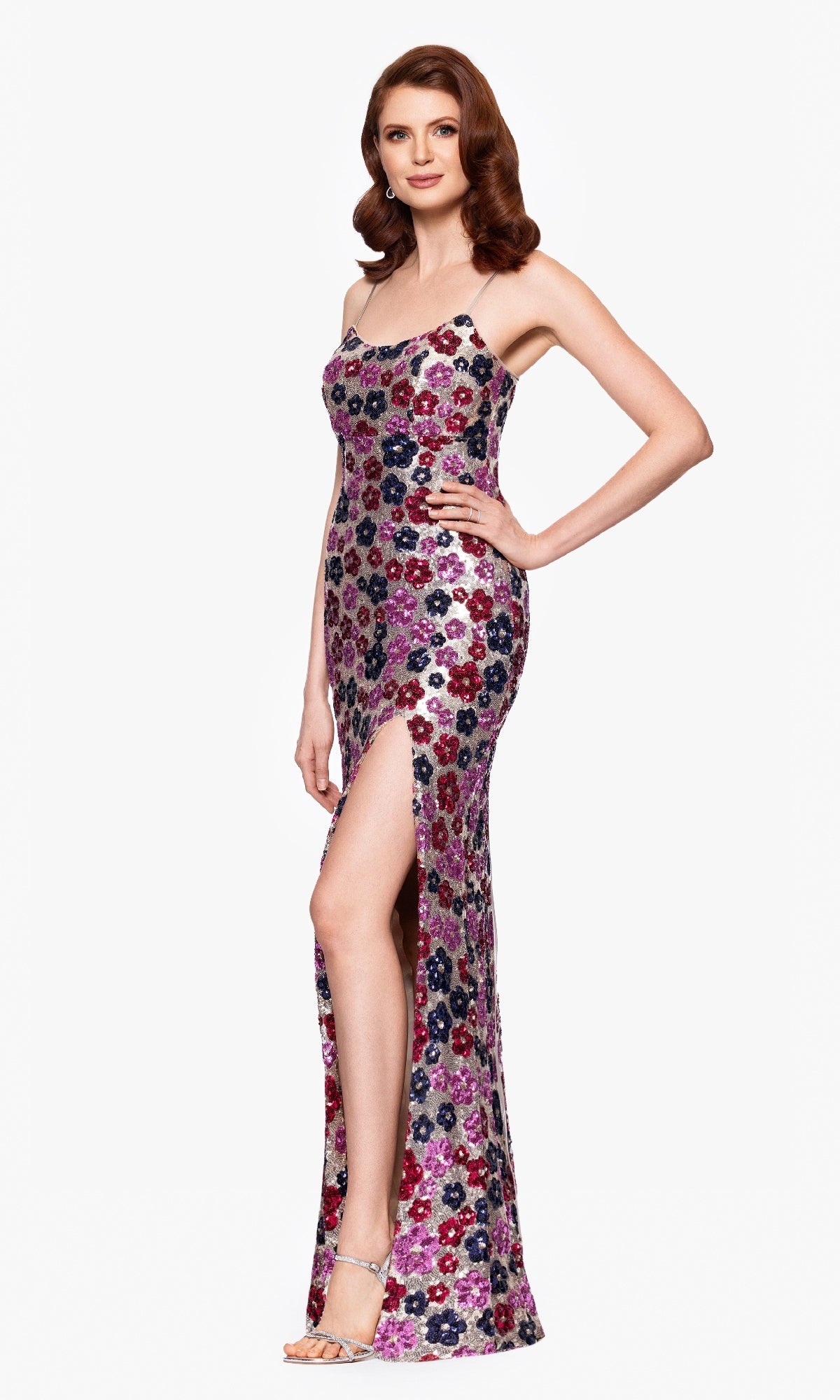A woman in the Betsy & Adam A27017 Floral-Sequin Long Prom Dress, featuring a sleeveless design, colorful patterns, and a thigh-high slit, stands smiling against a plain background.